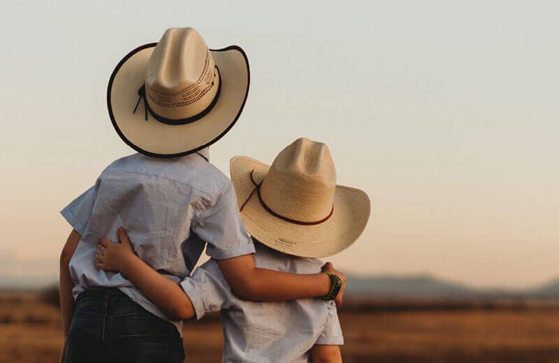 Two children in akubra hats looking out over a paddock at sunset